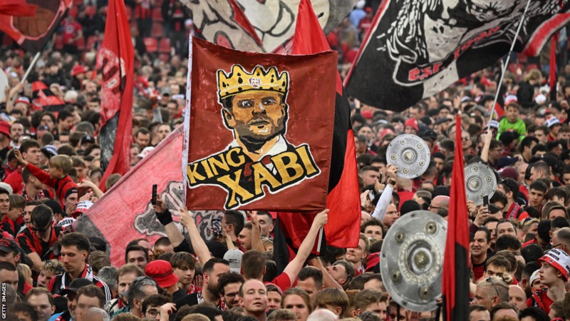 Pendukung Leverkusen yang membentangkan bendera bergambar Xabi Alonso sebagai tanda penghormatan (Sumber Foto : Getty Images) - Image