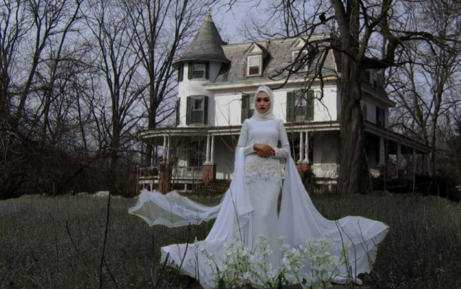 Seorang model mengenakan gaun putih dengan ekor panjang dan berhijab pocong berfoto dengan tatapan seram di depan sebuah gedung tua. - Image