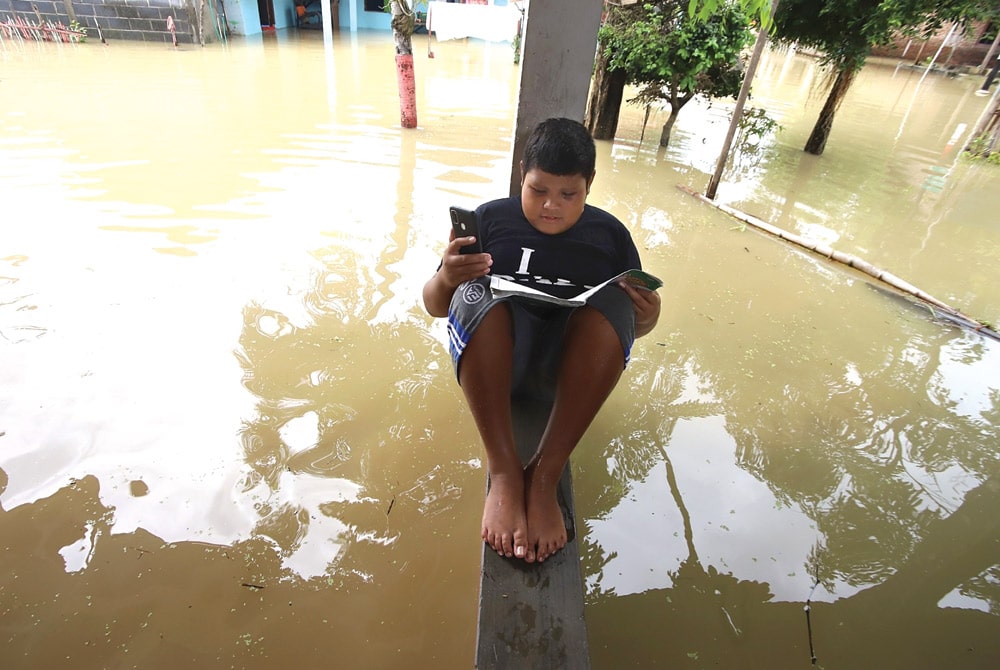 DI TENGAH AIR BAH: Aldo belajar secara daring di teras rumah di tengah banjir yang masih menggenangi Dusun Balong, Desa Banyulegi, Dawarbalndong, Mojokerto, kemarin (14/12). (SOFAN KURNIAWAN/JAWA POS RADAR MOJOKERTO) - Image